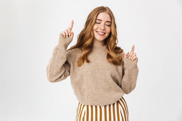 Beautiful young girl with long blond curly hair wearing sweater standing isolated over white wall, pointing at copy space