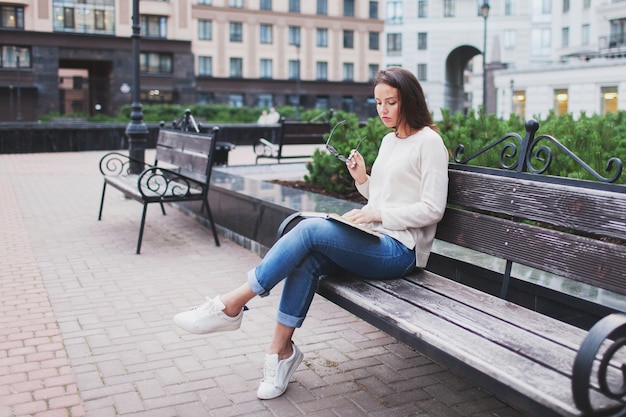 A beautiful young girl with long brown hair sitting on a bench with a book, holding eyeglasses