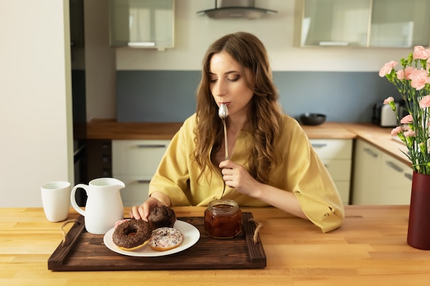 A beautiful young girl with long hair is sitting at home in the kitchen and is planning to have breakfast.