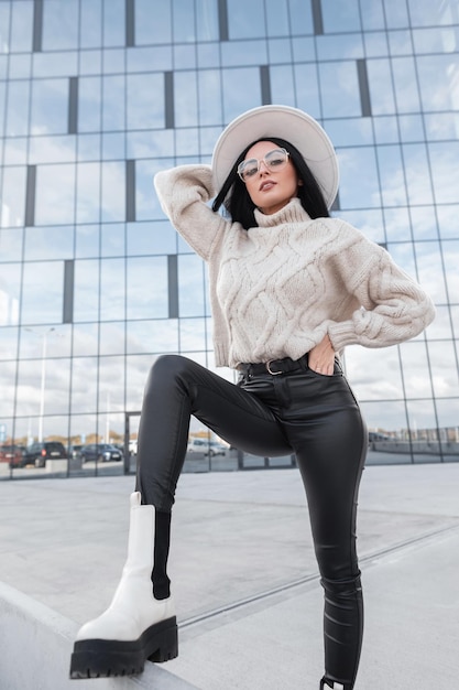 Beautiful young girl with long hair and stylish glasses in\
fashionable knit clothes with a hat leather pants and fashion white\
leather shoes poses in the city