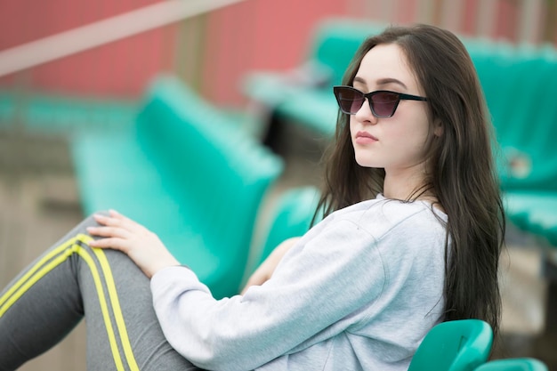 Beautiful young girl with long hair in sunglasses in the sports standsWoman in sport pants