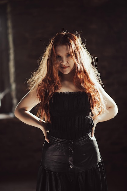 Beautiful young girl with long red hair in a black dress in a dark room