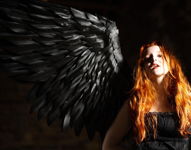 Beautiful young girl with long red hair and black wings in a dark room