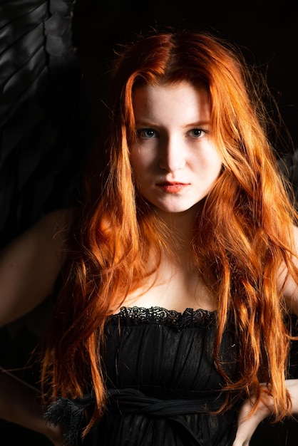 Beautiful young girl with long red hair and black wings in a dark room