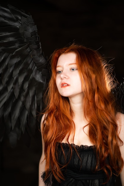 Beautiful young girl with long red hair and black wings in a dark room