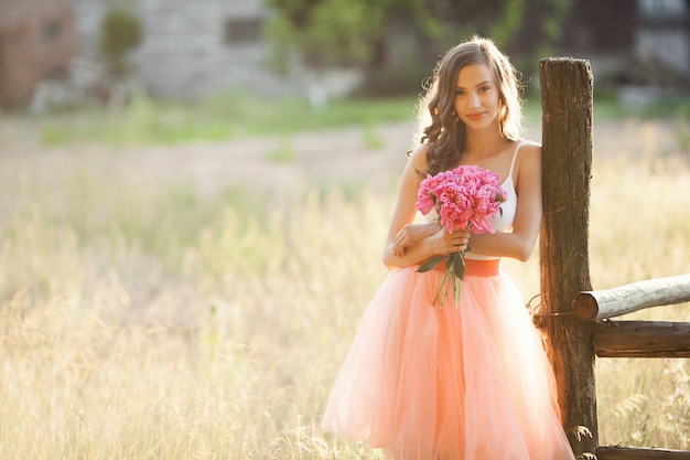 Beautiful young girl with pions on the nature outdoors in the sunshine. Pretty woman outside with flowers in the pink skirt