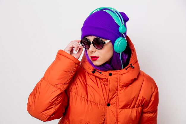 Красивая молодая девушка с фиолетовыми волосами и в оранжевой куртке слушает музыку в наушниках.