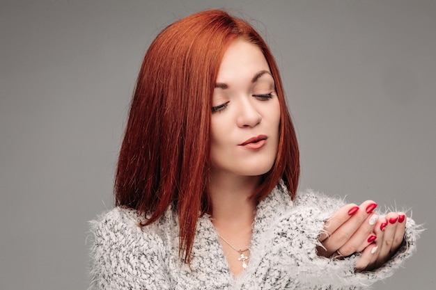 Beautiful young girl with red hair and red nails holding her hands together and blowing