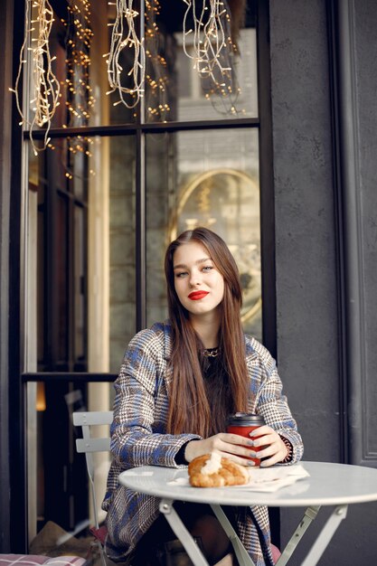 야외 카페에 앉아 빨간 머리를 가진 아름 다운 젊은 여자. 커피를 마시고 크루아상을 먹는 소녀. 소녀는 세련된 파란색 코트를 입고 빨간 입술을 가지고 있습니다.