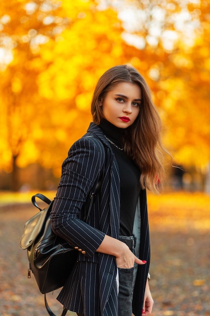 Beautiful young girl with red lips in a fashionable blazer with a black sweater, chain and backpack walks in nature with golden autumn foliage at sunset