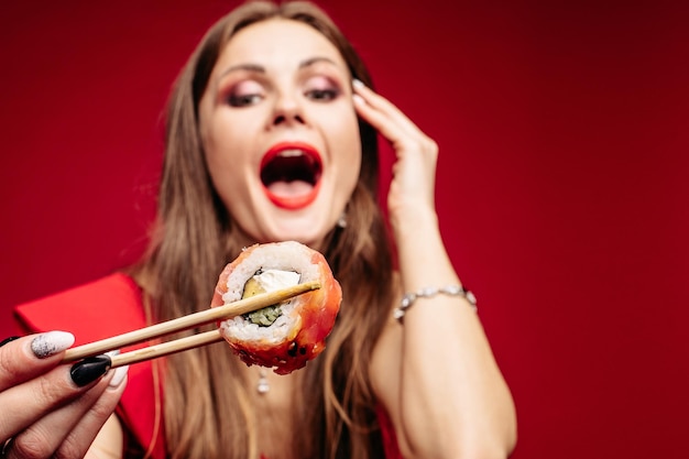 Beautiful young girl with red lips holding wooden sticks in her hand Brunette model with long hair eating asian food Pretty lady in red blouse with widely opened mouth enjoying her tasty meal