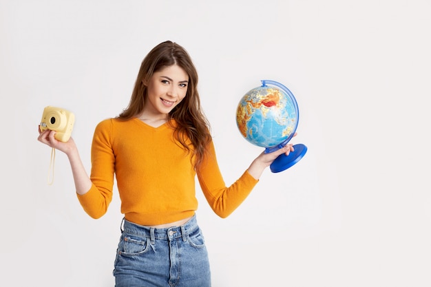 A beautiful young girl in a yellow sweater is holding a globe and a camera. Recreation, travel, tourism. Space for text