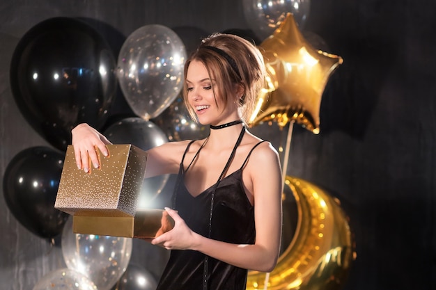 Красивая молодая великолепная белокурая женщина в темном черном вечернем платье открывает золотую подарочную коробку, на вечеринке, праздничной, праздничной черной предпосылке. Рождество или день рождения концепция с подарком в руках девушки, полная длина