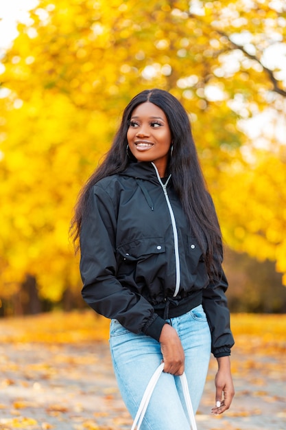 Beautiful young happy black canadian woman with a smile in a casual jacket with jeans walks in the park with a yellow fall leaves on autumn vacation