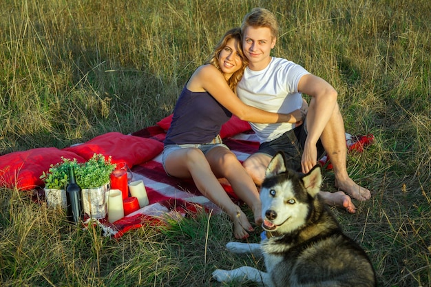 Красивая молодая счастливая любящая пара на пикнике, лежа на пледе с их собакой в поле в солнечный летний день, наслаждаясь и отдыхая. смотрит в камеру и улыбается.