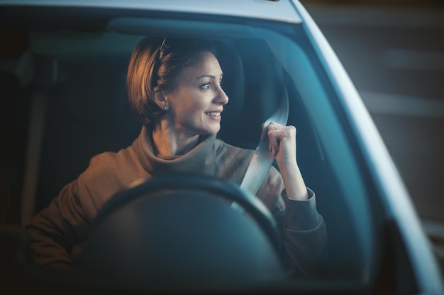 Beautiful young happy woman is sitting on car seat, fastening seat belt and looking through car window.