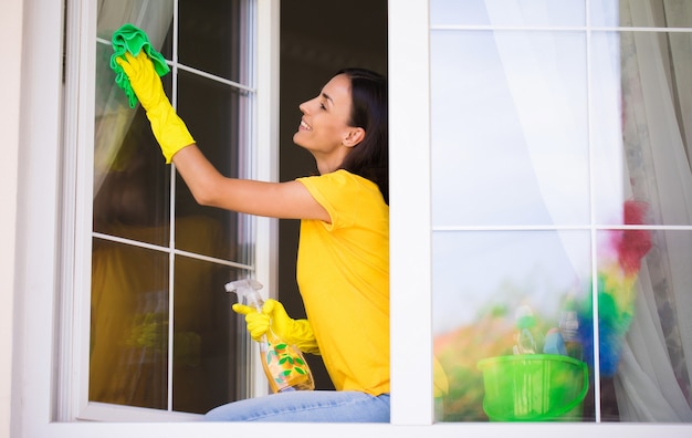 beautiful young happy woman is using a duster and a spray while cleaning a window in the house