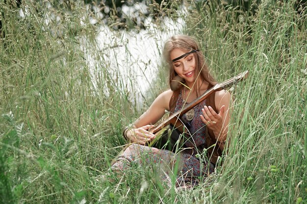 Beautiful young hippie woman playing guitar sitting on forest glade