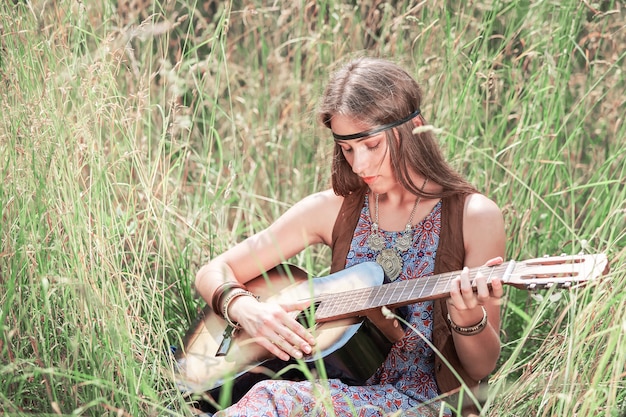 Bella giovane donna del hippie che gioca chitarra che si siede sull'erba