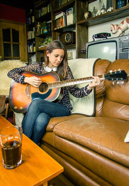 Photo beautiful young hipster girl playing acoustic guitar sitting in a sofa at home. retro vintage colors edition