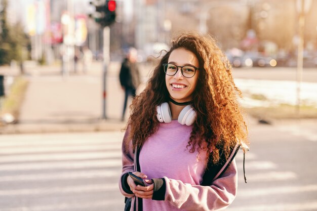 美しい若い流行に敏感な混血少女通りに立って、スマートフォンを押しながら探して