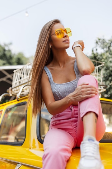 Premium Photo  Beautiful young hipster woman in fashionable summer clothes  with a sexy top pink pants sneakers and yellow glasses sits and rest on a  vintage car female summer colorful look