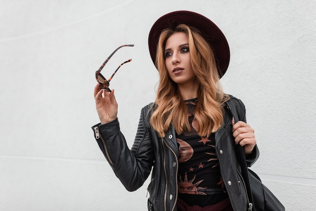 Beautiful young hipster woman with dark sunglasses in a trendy purple hat in a leather jacket with a black stylish backpack