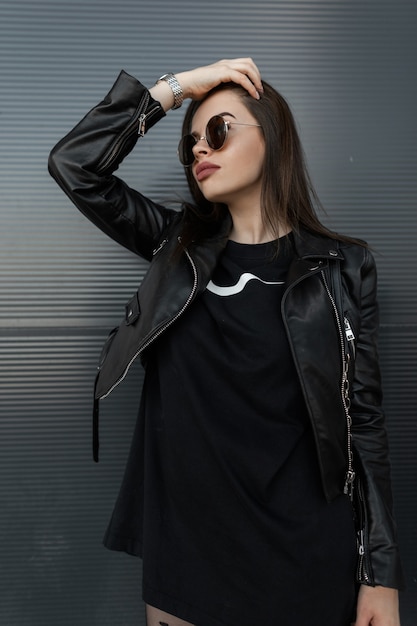 Bella giovane donna hipster con eleganti occhiali da sole rotondi in una giacca di pelle alla moda con un vestito nero si trova vicino a un muro di metallo sulla strada