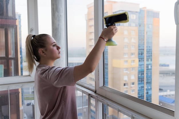 Beautiful young housewife in working clothes washing windows by professional tools at home.