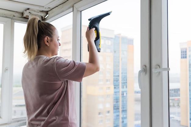 Beautiful young housewife in working clothes washing windows by professional tools at home.