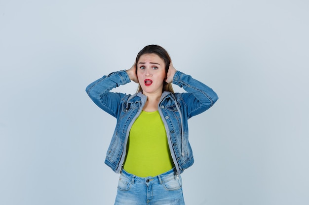 Beautiful young lady keeping hands on ears in denim outfit and looking scared , front view.