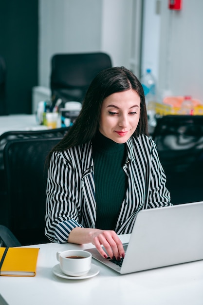 Beautiful young lady looking attentively at the laptop screen in a modern office and smiling