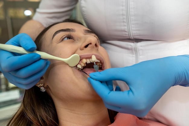 치과 진료소에 앉아 있는 아름다운 젊은 여성은 올바른 치아 색상을 미백하기 위해 치아 색상 샘플을 선택합니다.