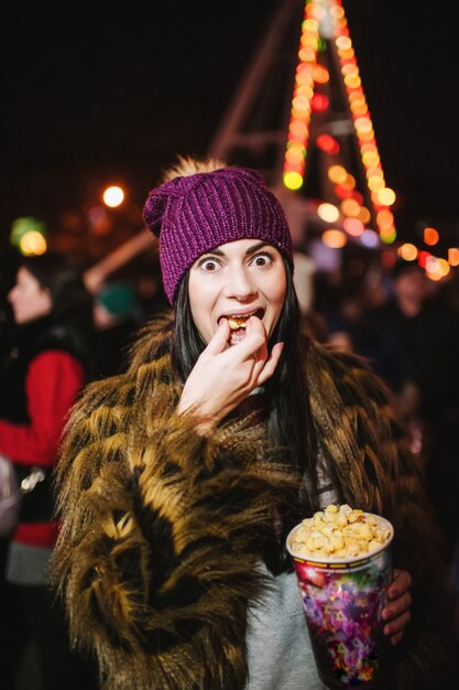 カルーセルの近くのクリスマスフェアに立って、ポップコーンを食べている美しい若い女性