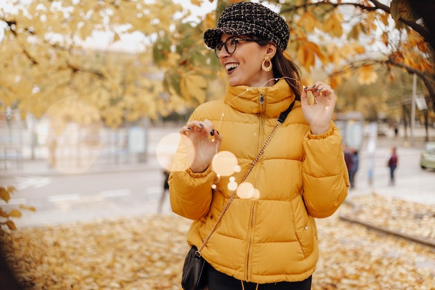 Foto bella giovane donna in abiti caldi eleganti sorridendo e toccando le foglie gialle sull'albero in un meraviglioso giorno d'autunno in una strada della città