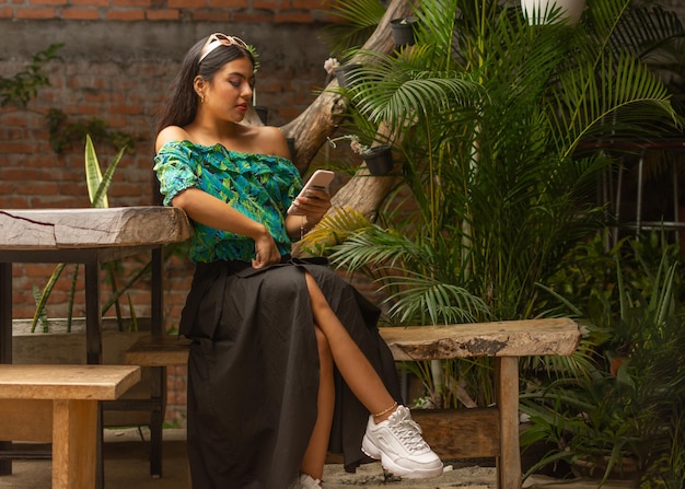 Красивая молодая латинская модная брюнетка с солнцезащитными очками на голове, небрежно одетая, смотрит на свой смартфон, сидя на деревенской скамейке с натуральными растениями на заднем плане