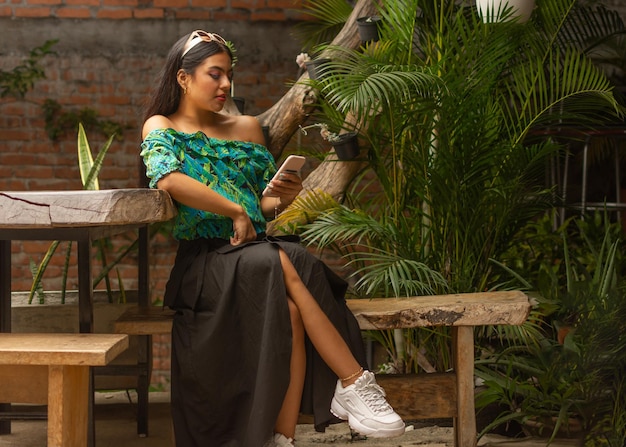 Beautiful young latin fashionist brunette with sunglasses over head casually dressed looking her smart phone sitting on a rustic bench with natural plants in background