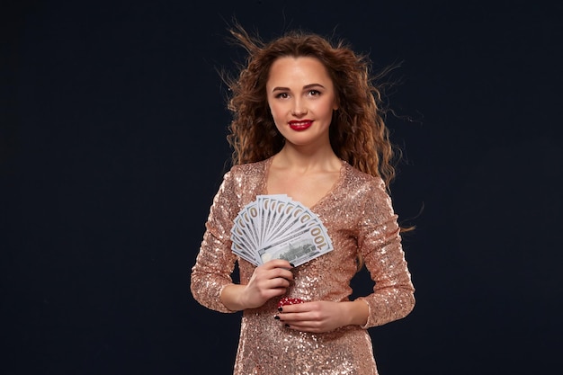 Beautiful young lucky happy brown-haired woman in golden cocktail dress showing the money fan in one hand and poker chips in another hand, fan of 100 dollar bills, black background. Gambling, casino,