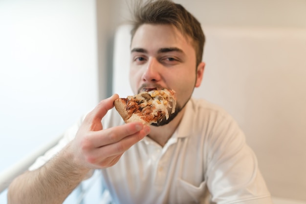 美しい若い男が食欲をそそるピザを食べ、カメラを見ます。ひげの男の子がピザを一皿料理します。