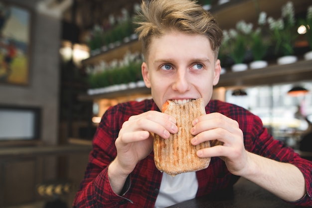 A beautiful young man eats a panini sandwich in a cozy cafe.