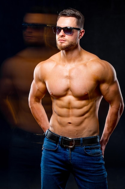 Beautiful young man with perfect body posing on dark background Strong hands in back pockets Guy in sunglasses Dubling reflection Studio photoshoot