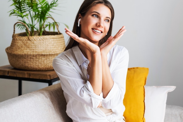 Beautiful young millennial girl is sitting in relaxed position on sofa. Woman is resting after hard day. The girl in white shirt is very happy to be at home. 