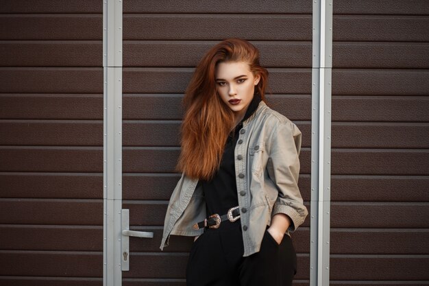 Beautiful young model girl in stylish jacket and black polo shirt posing near the gate