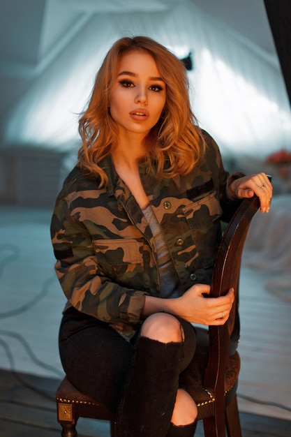 Красивая молодая модель женщина в модной военной куртке сидит на стуле в студии