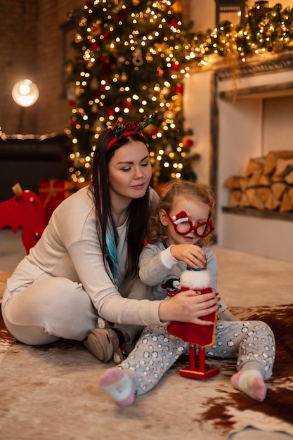 Beautiful young mom and fun daughter girl child in fashionable pajamas clothes sit and play with a toy near the fireplace with Christmas decorations and lights
