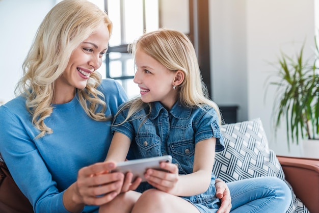 Beautiful young mom and her little daughter using smartphone and smiling while sitting on sofa