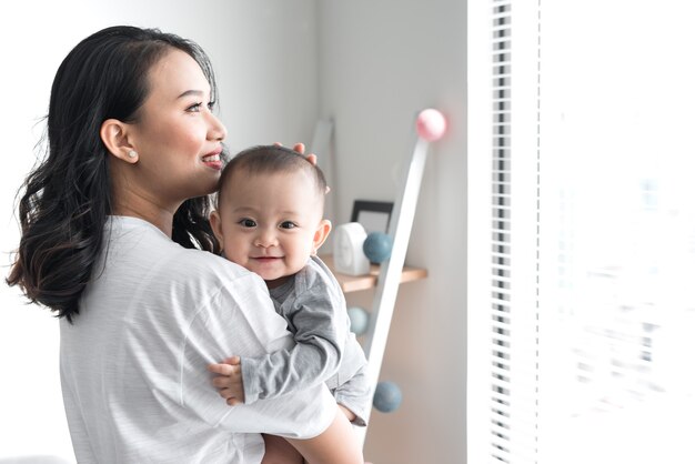 Photo beautiful young mom is playing with her cute baby and smiling while standing near the window at home