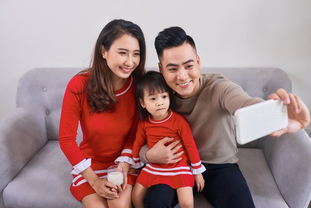 Beautiful young mother, father and their daughter are making selfie using a phone and smiling while sitting on the sofa at home