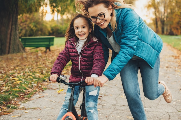 Bella giovane madre che aiuta a sua figlia ad imparare ad andare in bicicletta nel parco. bambina sveglia che impara ad andare in bicicletta con sua madre.