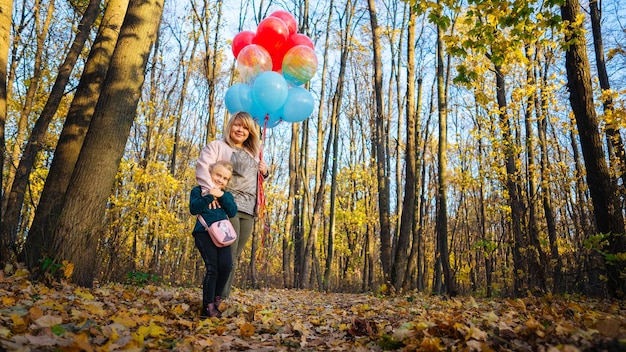 A beautiful young mother hugs her a little adorable daughter and holds the colorful balloons on a wall walk in autumn forest